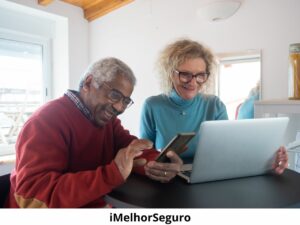 um homem de vermelho no celular e uma mulher de azul no notebook. Ambos usam óculos, estão em uma residência, sentados à mesa e sorrindo