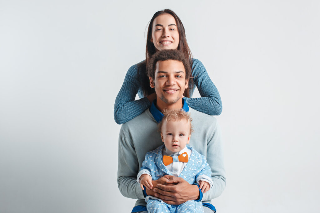 família de 3 pessoas se abraçando: uma mulher jovem e um homem jovem, ambos de blusa de frio, além de um bebê no colo do homem, usando um macacão azul pastel